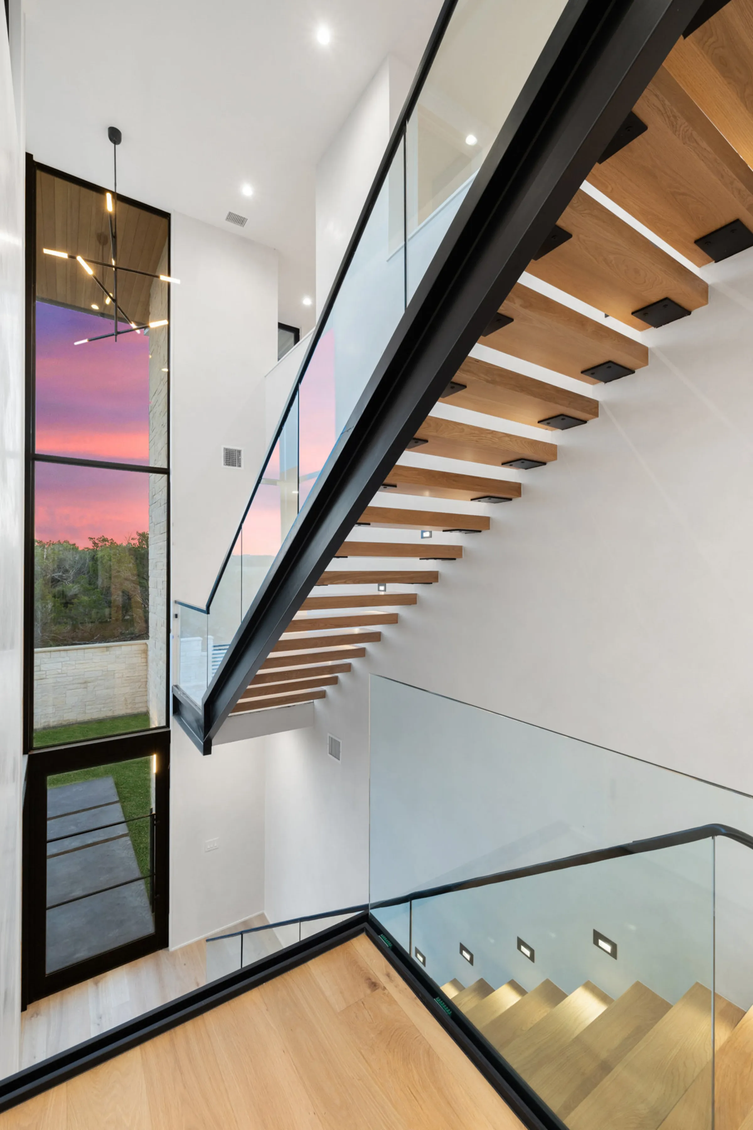 A floating oak staircase against sunset glass — Seven Custom Homes